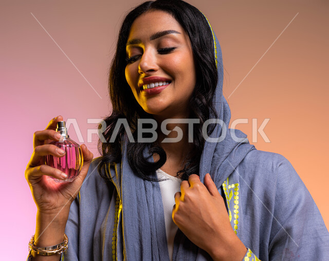 إيماءات الإعجاب بالروائح العطرة, التسويق لمنتجات العطور والمستحضرات النسائيه، مفهوم الأناقة والإهتمام بالمظهر الخارجي، بورتريه مقرب لإمرأه عربيه سعودية خليجية مبتسمة تقوم بالتطيب والتعطر بالعطور النسائيه الخليجيه والزيوت العطرية، خلفية ملونه