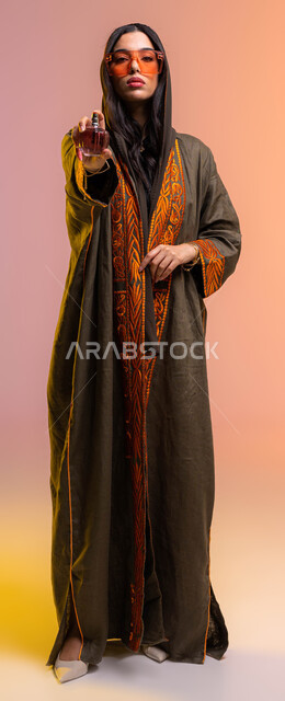 مفهوم الأناقة والإهتمام بالمظهر الخارجي, بورتريه لإمرأة عربية سعودية خليجية تقوم برش العطر, التسويق لمنتجات العطور والمستحضرات النسائية, التطيب والتعطر بالعود والزيوت العطرية. ارتداء العباءة والنظارات الشمسية, صورة الجسم بالطول الكامل, خلفية ملونة