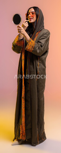 مفهوم الإهتمام بالجمال والبشرة, أدوات مكياج تتوريال, التسويق لمستحضرات التجميل في المتاجر و عبر الانترنت, بورتريه لخبيرة تجميل عربية خليجية تستخدم إحدى أدوات الميكآب(أحمر الشفاه)وتنظر إلى المرآة, ارتداء النظارة الشمسية, صورة الجسم بالطول الكامل, خلفية ملو