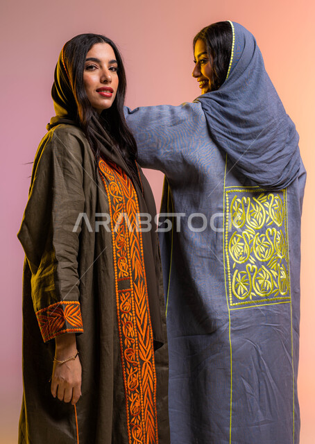 عرض الأزياء والموديلات الحديثة, مفهوم الموضة والأناقة, عباءات خليجية نسائية عصرية, بورتريه لصديقتان عربيتان سعوديتان خليجيتان ترتديان عبايتان ملونة أنيقة, ايماءات تدل على الصداقه والمحبة, شراء الملابس والاكسسوارات النسائية, خلفية ملونة