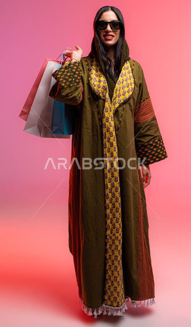 International and Saudi brands, the pleasure of shopping in malls, a portrait of a Saudi Arabian Gulf woman wearing an elegant abaya holding colorful shopping bags in her hand, seasonal offers and discounts, wearing sunglasses, beauty and femininity gestu