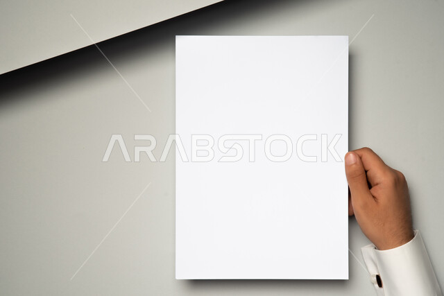 A close-up of the hand of a Saudi Arabian Gulf man holding white papers, a mockup of white paper on the table, office supplies and stationery