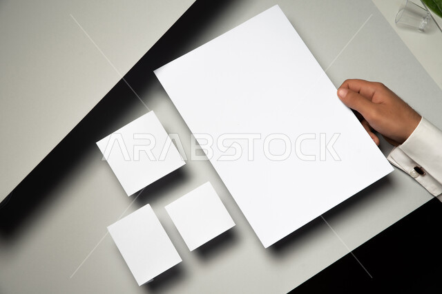 موك اب لمجموعة من اوراق ملاحظات صغيرة على الطاولة، صورة مقربة ليد رجل عربي سعودي خليجي يمسك بورق ابيض، ادوات وقرطاسية مكتبية