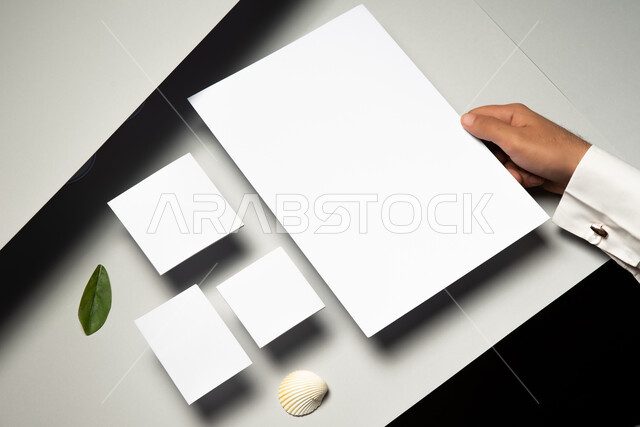 ادوات وقرطاسية مكتبية، موك اب لمجموعة من اوراق ملاحظات صغيرة على الطاولة، صورة مقربة ليد رجل عربي سعودي خليجي يمسك بورق ابيض، ورقة شجر خضراء