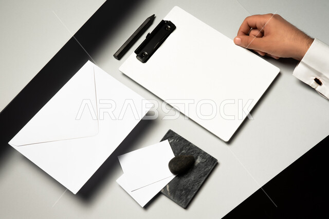 اوراق ملاحظات صغيرة، ادوات وقرطاسية مكتبية، صورة مقربة ليد رجل عربي سعودي خليجي يمسك بملف ابيض، موك اب لمظروف رسائل على الطاولة، ورقة بيضاء