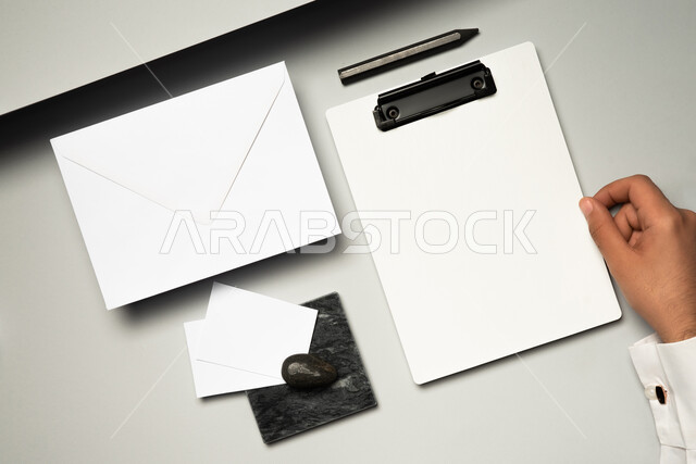 موك اب لمظروف رسائل على الطاولة، صورة مقربة ليد رجل عربي سعودي خليجي يمسك بملف ابيض، اوراق ملاحظات صغيرة، ادوات وقرطاسية مكتبية، ورقة بيضاء