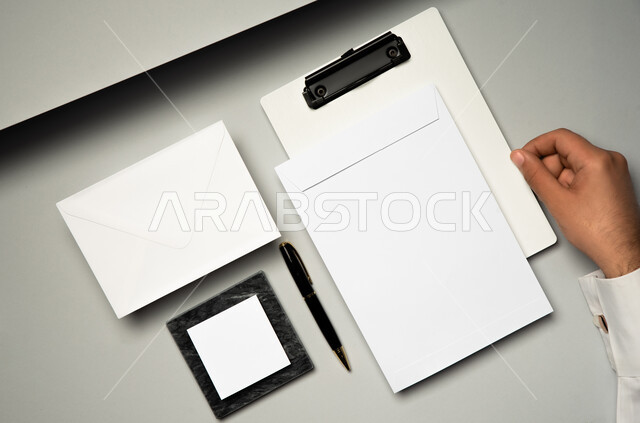 اوراق ملاحظات صغيرة، صورة مقربة ليد رجل عربي سعودي خليجي يمسك بملف ابيض، ادوات وقرطاسية مكتبية، موك اب لمظروف رسائل على الطاولة، ورقة بيضاء