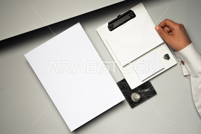 اوراق ملاحظات صغيرة، صورة مقربة ليد رجل عربي سعودي خليجي يمسك ملف فوقه مظروف رسائل، ادوات وقرطاسية مكتبية، موك اب لورق ابيض على الطاولة، ورقة بيضاء