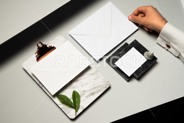 ورقة ملاحظات صغيرة، ادوات وقرطاسية مكتبية، موك اب لورق ابيض على الطاولة، صورة مقربة ليد رجل عربي سعودي خليجي يمسك بمظروف رسائل، اوراق الاشجار الخضراء