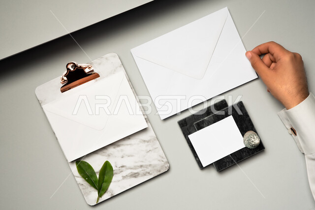 Green leaves, office supplies and stationery, mockup of white paper on the table, close-up of the hand of a Saudi Arabian Gulf man holding an envelope of letters, a small paper of notes