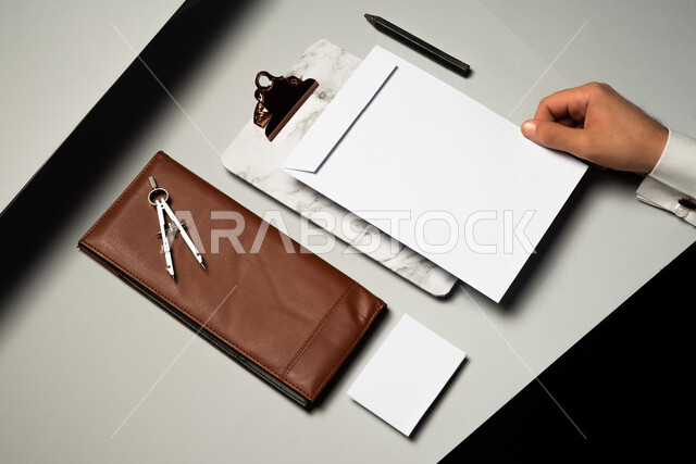 اوراق ملاحظات صغيرة، أدوات وقرطاسية مكتبية، موك اب لمحفظة شخصية على الطاولة، صورة مقربة ليد رجل عربي سعودي خليجي يمسك بمظروف رسائل، ورقة بيضاء