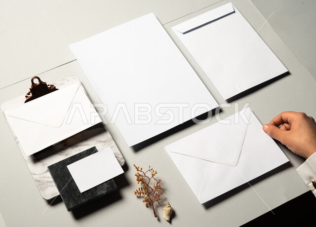 موك اب لورق ابيض على الطاولة، صورة مقربة ليد رجل عربي سعودي خليجي يمسك بمظروف رسائل، اوراق ملاحظات صغيرة، أدوات وقرطاسية مكتبية، ورقة بيضاء