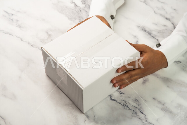 خلفية رخام ابيض، صورة مقربة ليد رجل سعودي عربي خليجي يحمل صندوق ابيض، كراتين الشحن والتوصيل الاونلاين، تسليم الطلبات والبضائع للزبائن، موك اب لصندوق كرتوني مغلف على طاولة رخامية بيضاء، صناديق كرتون تعبئة وتغليف، خدمات التوصيل السريع والامن