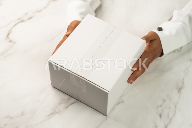خدمات التوصيل السريع والامن، موك اب لصندوق كرتوني مغلف على طاولة رخامية بيضاء، صورة مقربة ليد رجل سعودي خليجي عربي يمسك صندوق ابيض، كراتين الشحن والتوصيل الاونلاين، تسليم البضائع والطلبات للزبائن، صناديق كرتون تعبئة وتغليف، خلفية رخام ابيض