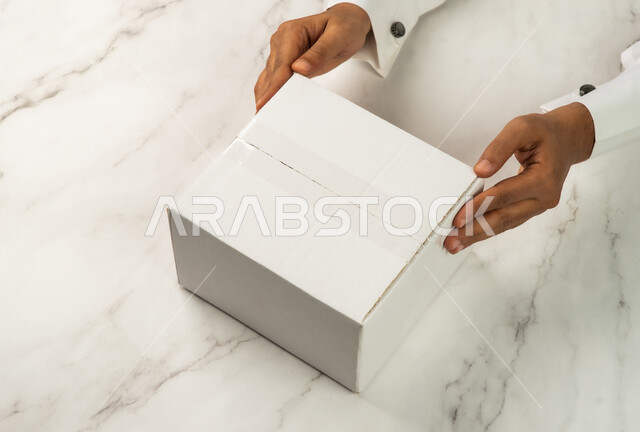 خلفية رخام ابيض، خدمات التوصيل السريع والامن، صورة مقربة ليد رجل سعودي خليجي عربي يحمل صندوق ابيض، موك اب لصندوق كرتوني مغلف على طاولة رخامية بيضاء، كراتين الشحن والتوصيل الاونلاين، تسليم البضائع والطلبات للزبائن، صناديق كرتون تعبئة وتغليف