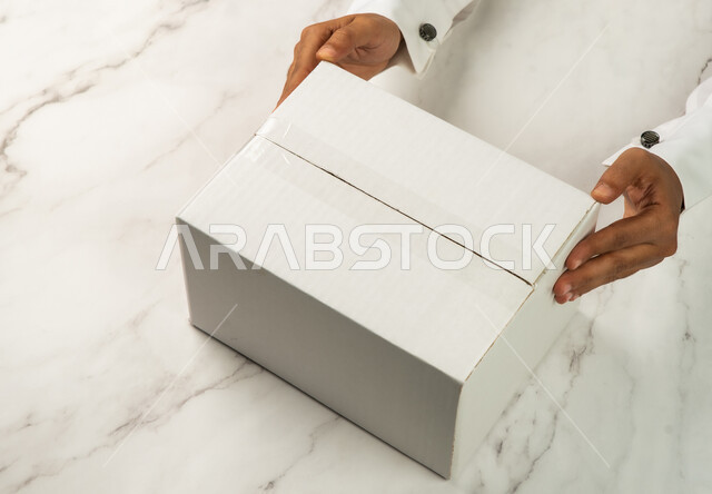 خدمات التوصيل السريع والامن، صورة مقربة ليد رجل سعودي عربي خليجي يحمل صندوق ابيض، موك اب لصندوق كرتوني مغلف على طاولة رخامية بيضاء، كراتين الشحن والتوصيل الاونلاين، تسليم الطلبات والبضائع للزبائن، صناديق كرتون تعبئة وتغليف، خلفية رخام ابيض