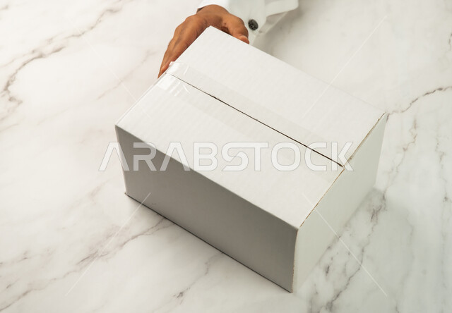 صناديق كرتون تعبئة وتغليف، صورة مقربة ليد عربي رجل سعودي خليجي يحمل صندوق ابيض، موك اب لصندوق كرتوني مغلف على طاولة رخامية بيضاء، كراتين الشحن والتوصيل الاونلاين، تسليم البضائع والطلبات للزبائن، خدمات التوصيل السريع والامن، خلفية رخام ابيض