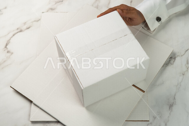 Mockup of a wrapped carton box on a white marble table, a close-up of a Saudi Arabian Gulf man's hand holding a white box, online shipping and delivery cartons, cardboard packaging boxes, express and security delivery services, delivery of orders and good