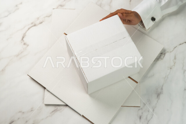 تسليم الطلبات والبضائع للزبائن، موك اب لصندوق كرتوني مغلف على طاولة رخامية بيضاء، صورة مقربة ليد رجل عربي خليجي سعودي يحمل صندوق ابيض، كراتين الشحن والتوصيل الاونلاين، صناديق كرتون تعبئة وتغليف، خدمات التوصيل السريع والامن، خلفية رخام ابيض