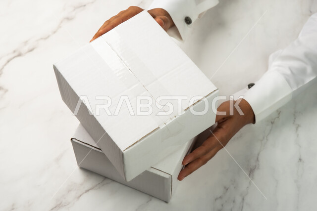 كراتين الشحن والتوصيل الاونلاين، صورة مقربة ليد رجل سعودي عربي خليجي يحمل صندوق ابيض، موك اب لصندوق كرتوني مغلف على طاولة رخامية بيضاء، تسليم البضائع والطلبات للزبائن، صناديق كرتون تعبئة وتغليف، خدمات التوصيل السريع والامن، خلفية رخام ابيض