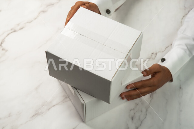 صورة مقربة ليد رجل عربي سعودي خليجي يحمل صندوق ابيض، موك اب لصندوق كرتوني مغلف على طاولة رخامية بيضاء، كراتين الشحن والتوصيل الاونلاين، تسليم الطلبات والبضائع للزبائن، صناديق كرتون تعبئة وتغليف، خدمات التوصيل السريع والامن، خلفية رخام ابيض