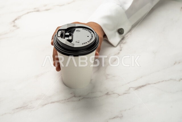 خلفية رخام ابيض، موك اب لكوب من الورق المقوى على طاولة رخامية بيضاء، شراء القهوة الطازجة من المقاهي السعودية، صورة مقربة ليد رجل عربي خليجي سعودي يمسك كوب القهوة، خدمات التوصيل السريع والامن، تسليم المشتريات والطلبات للزبائن