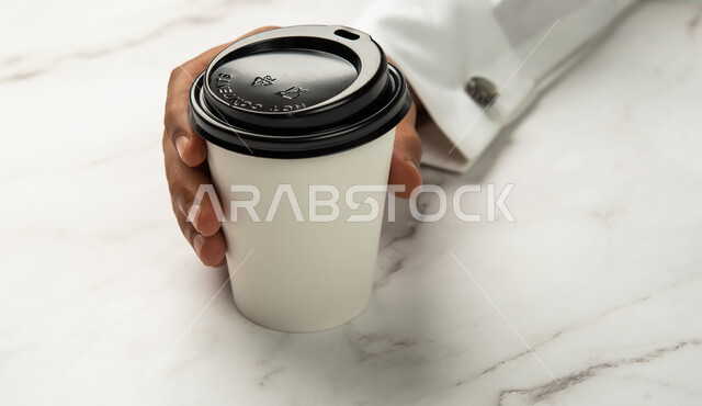 موك اب لكوب من الورق المقوى على طاولة رخامية بيضاء، خلفية رخام ابيض، شراء القهوة الطازجة من المقاهي السعودية، صورة مقربة ليد رجل عربي خليجي سعودي يمسك كوب القهوة، خدمات التوصيل السريع والامن، تسليم المشتريات والطلبات للزبائن