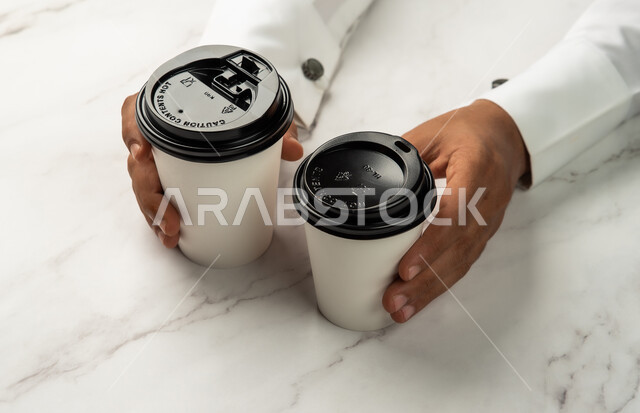 خدمات التوصيل السريع والامن، موك اب لاكواب من الورق المقوى على طاولة رخامية بيضاء، شراء القهوة الطازجة من المقاهي السعودية، صورة مقربة ليد رجل عربي خليجي سعودي يمسك كوب القهوة، تسليم المشتريات والطلبات للزبائن، خلفية رخام ابيض