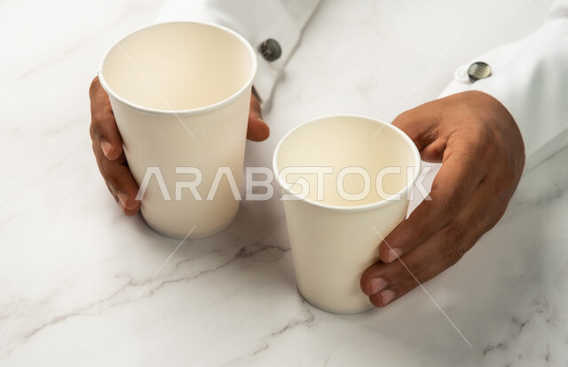 شراء القهوة الطازجة من المقاهي السعودية، موك اب لاكواب من الورق المقوى على طاولة رخامية بيضاء، صورة مقربة ليد رجل عربي خليجي سعودي يمسك كوب القهوة، خدمات التوصيل السريع والامن، تسليم المشتريات والطلبات للزبائن، خلفية رخام ابيض
