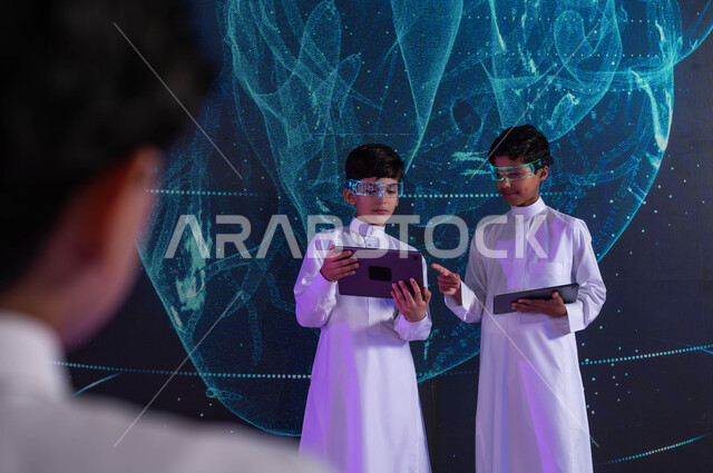 دعم التطوير التكنولوجي والتجديد والابداع في المؤسسات التربوية، طالبان سعوديان عربيان خليجيان يرتديان نظارات الواقع الافتراضي ويستخدمان اجهزة التابليت، خلفية شاشات هولوجرامية ثلاثية الأبعاد لقلب الإنسان، رؤية مستقبلية 2030