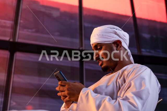 Waiting to board the plane at sunset, a close-up photo of an Egyptian tourist wearing a galabiya and turban sitting in the waiting lounge at the airport, using a mobile phone to chat and socialize, spending an enjoyable summer trip in Saudi Arabia, arriving and receiving tourists from abroad