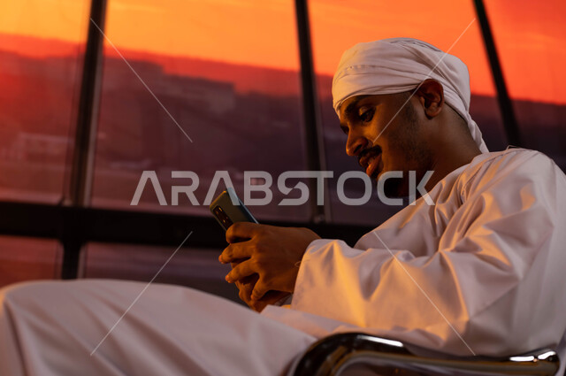 قضاء رحلة صيفيه ممتعة في السعوديه، إنتظار ركوب الطائرة عند غروب الشمس، صورة مقربه لسائح مصري يرتدي الجلابية و العمامة يجلس في صاله الإنتظار في المطار يستخدم الهاتف المحمول للدردشة و التواصل الإجتماعي، قدوم و استقبال السياح من الخارج