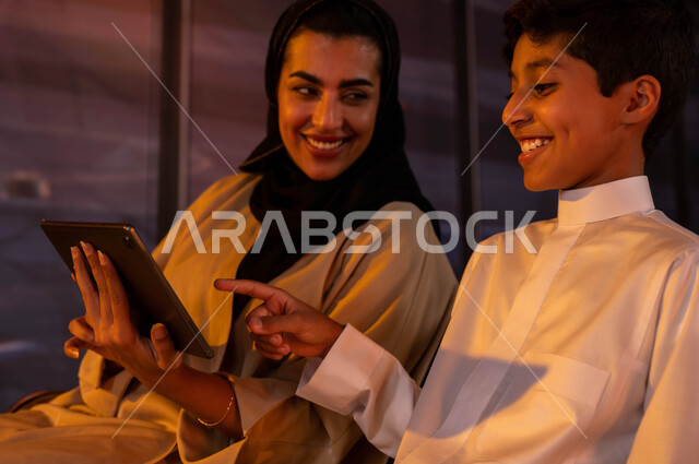 مشاهدة البرامج الترفيهية و اللعب مع الأبناء عبر جهاز التابلت أثناء إنتظار ركوب الطائره، أم عربية خليجية سعودية تجلس مع إبنها في صالة المطار،  إيماءات السرور والاستمتاع مع الأطفال, الاستعداد  للسفر و السياحة، إجازه عائليه صيفيه ممتعة, رحلات جويه سعودية