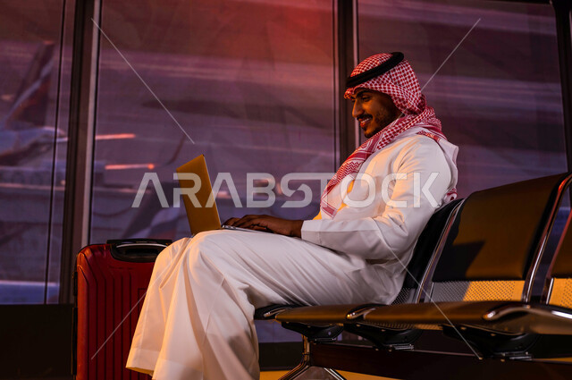  أهمية السياحة في السعودية, إنجاز العمل من خلال جهاز الحاسوب, إقامة حجوزات فندقية الكترونية وتصفح الانترنت ومتابعة الأخبار, رجل أعمال عربي سعودي خليجي يرتدي الثوب السعودي داخل صالة الانتظار في المطار وبجانبه حقيبة السفر, الاستعداد للسفر في رحلة عمل