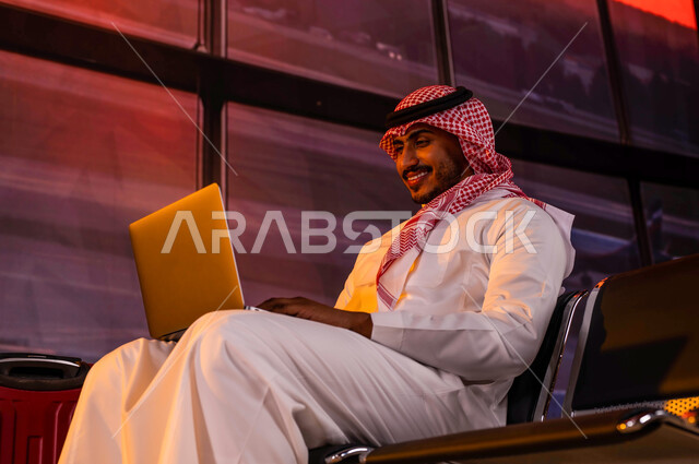 إنجاز العمل من خلال جهاز الحاسوب, إقامة حجوزات فندقية وتصفح الانترنت ومتابعة الأخبار, أهمية السياحة في السعودية, رجل أعمال عربي سعودي خليجي يرتدي الثوب السعودي التقليدي داخل صالة الانتظار في المطار وبجانبه حقيبة السفر, الاستعداد للسفر في رحلة عمل