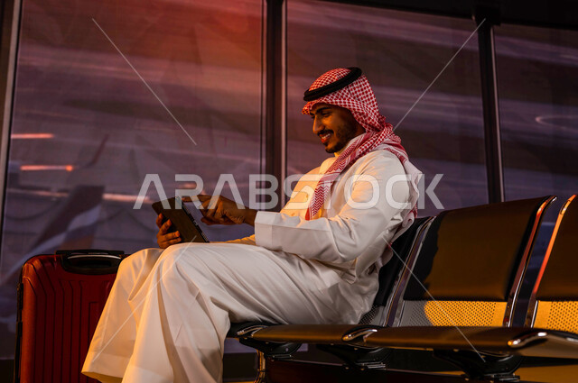 إقامة حجوزات فندقية الكترونية ومشاركة تفاصيل الرحلة على مواقع التواصل الاجتماعي, تصفح الانترنت ومتابعة الأخبار, رجل أعمال عربي سعودي خليجي مبتسم يرتدي الثوب السعودي داخل قاعة الانتظار في المطار يقوم بإنجاز مهام العمل أثناء السفر عبر جهاز التابليت