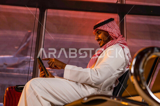قضاء إجازة صيفية ممتعة, إقامة حجوزات فندقية الكترونية, مشاركة تفاصيل الرحلة على مواقع التواصل الاجتماعي, تصفح الانترنت ومتابعة الأخبار, رجل أعمال عربي سعودي خليجي مبتسم يرتدي الثوب السعودي داخل قاعة الانتظار في المطار يقوم بإنجاز مهام العمل أثناء السفر