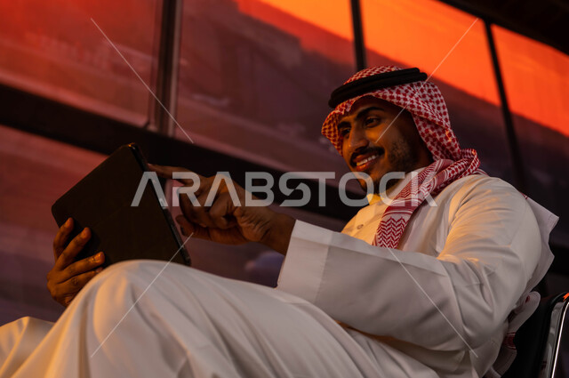 مشاركة تفاصيل الرحلة على مواقع التواصل الاجتماعي وتصفح الانترنت ومتابعة الأخبار, إقامة حجوزات فندقية الكترونية, رجل أعمال عربي سعودي خليجي مبتسم يرتدي الثوب السعودي داخل قاعة الانتظار في المطار يقوم بإنجاز مهام العمل عن بعد أثناء السفر عبر جهاز التابليت
