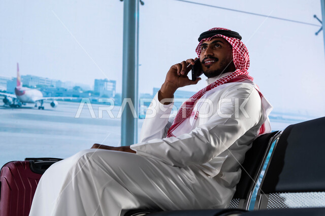 رحلة عمل جوية, رجل أعمال عربي سعودي خليجي مبتسم يرتدي الشماغ والثوب السعودي يقوم بإجراء مكالمة هاتفيه عبر الجوال, محادثات وتواصل اجتماعي, الجلوس في قاعة المطار والقيام بمناقشات العمل عبر الهاتف المحمول, الانتظار لركوب الطائرة والاستعداد للسفر والسياحة