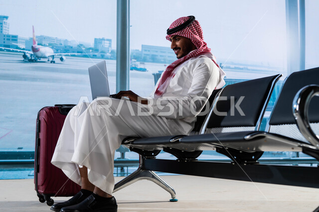 إقامة حجوزات فندقية الكترونية, تصفح الانترنت ومتابعة الأخبار, أهمية السياحة في السعودية, رجل أعمال عربي سعودي خليجي يرتدي الثوب السعودي داخل صالة الانتظار في المطار وبجانبه حقيبة السفر, إنجاز العمل من خلال جهاز الحاسوب, الاستعداد للسفر في رحلة عمل