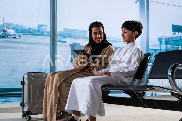 Gestures of joy at spending the holiday with the family, sharing flight details on social networking sites, Saudi Airlines, getting ready to go on a foreign tourist vacation, a Saudi Gulf Arab woman wearing a colorful abaya holding an iPad with her son at