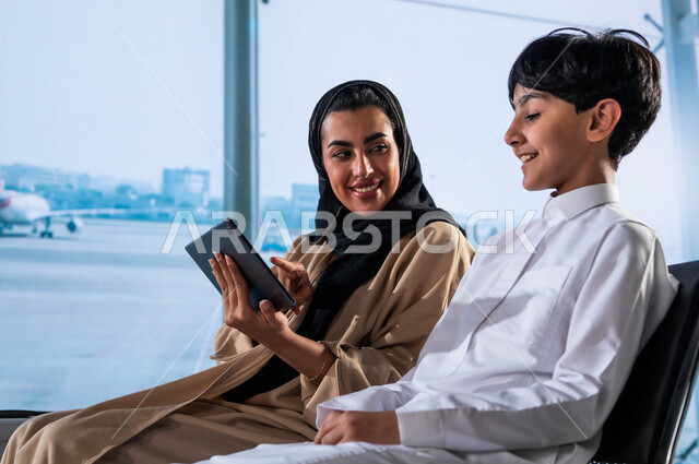 متعة السفر الجوي والسياحة العائلية الصيفية, الدردشة واللعب مع الأبناء على جهاز التابليت في قاعة الانتظار, ولد عربي سعودي خليجي يرتدي الثوب السعودي التقليدي يجلس مع والدته في المطار, تخطيط ومشاركة تفاصيل الرحلة مع العائلة, الاستعداد لركوب الطائرة