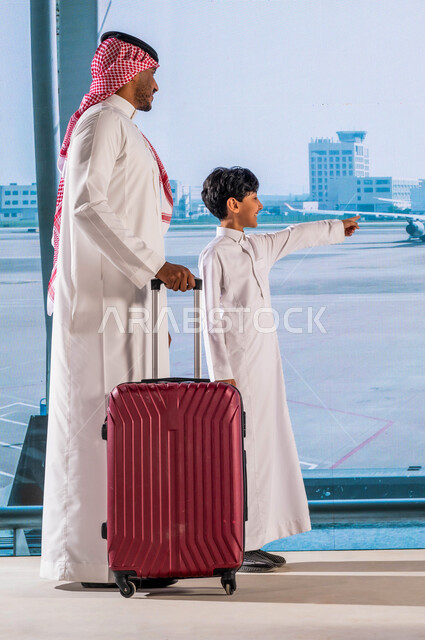 الاستعداد للذهاب في رحلة سياحية خارجية, قضاء أوقات عائلية ممتعة, رجل عربي خليجي سعودي يرتدي الشماغ والثوب السعودي التقليدي يمسك بيده حقيبة سفر برفقة ابنه في المطار, ايماءات يد تدل على الإشارة إلى الطائرة, صورة الجسم بالطول الكامل