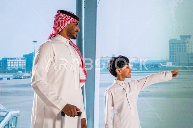 الاستعداد للسياحة والسفر في رحلة صيفية ترفيهية, ايماءات السعادة والسرور بقضاء أوقات عائلية ممتعة، رجل عربي خليجي سعودي يرتدي الشماغ والثوب السعودي التقليدي يمسك بيده حقيبة سفر برفقة إبنه في المطار, الإشارة والنظر إلى الطائرة من خلال النافذة