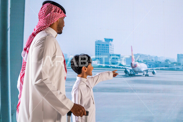 الإشارة والنظر إلى الطائرة من خلال النافذة, ايماءات السعادة والسرور بقضاء أوقات عائلية ممتعه، الاستعداد للسياحة والسفر في رحلة صيفية ترفيهية, رجل عربي خليجي سعودي يرتدي الشماغ والثوب السعودي التقليدي يمسك بيده حقيبة سفر برفقة إبنه في المطار