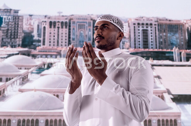 العمارة الإسلامية العريقة لقباب المساجد في المدينة المنورة، فضل الصلاة والدعاء في مسجد نبي الله، صورة مقربة لرجل سعودي عربي خليجي يرتدي الثوب السعودي التقليدي ويقف أمام المسجد النبوي الشريف ويدعو الله تعالى، خلفية القبب لمسجد النبي محمد صلى الله عليه وسلم
