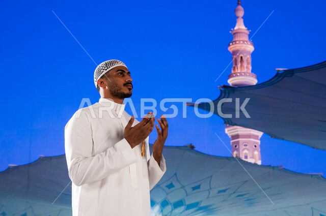  معالم و أماكن إسلامية مقدسه في السعوديه، صورة مقربه  لشاب مسلم عربي سعودي خليجي يحمل بيده سبحه و يدعو الله في ساحة المسجد النبوي الشريف في المدينة المنورة، الصلاة و الدعاء و العبادة وذكر الله تعالى، خلفية المئذنة و المظلات الإلكترونيه في مسجد رسول الله