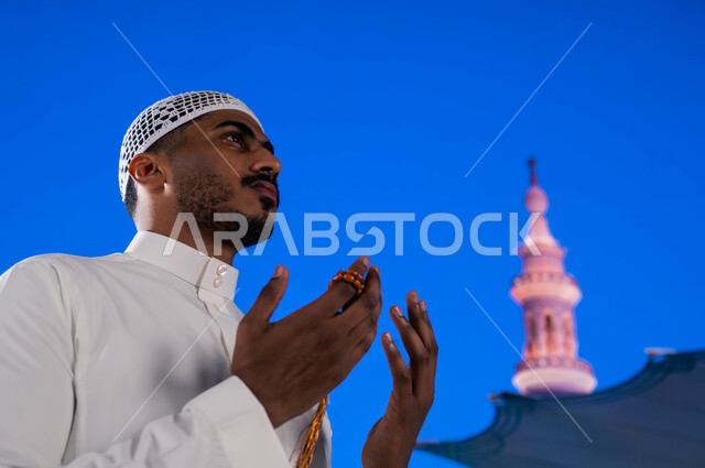 إيماءات الخشوع و التضرع إلى الله، أماكن و معالم إسلامية مقدسه في السعوديه، صورة مقربه  لشاب مسلم عربي سعودي خليجي يحمل بيده سبحه و يدعو الله في ساحة المسجد النبوي الشريف في المدينة المنورة، الصلاة و الدعاء و وذكر الله تعالى، خلفية المئذنة و المظلات