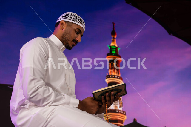 إيماءات تفسير وتدبر القرآن الكريم، فضل الصلاة و الدعاء في المسجد النبوي الشريف، قدوم المسلمين من شتى أنحاء العالم الى الأماكن المقدسه في المدينة المنورة، رجل عربي سعودي يقوم بقراءة وتلاوة القران، التقرب إلى الله، خلفية المئذنة و المظلات الإلكترونية