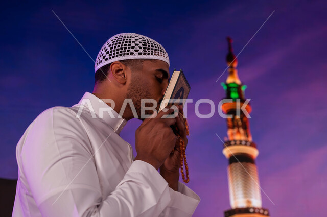 إيماءات تقبيل القرآن الكريم، فضل الصلاة و الدعاء في المسجد النبوي الشريف، قدوم المسلمين من شتى أنحاء العالم إلى الأماكن المقدسه في المدينة المنورة، رجل مسلم عربي سعودي يقوم بقراءة القرآن،  العبادة و التقرب إلى الله، خلفية المئذنة و المظلات الإلكترونيه